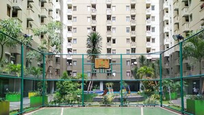 An outdoor basketball court surrounded by a green fence, with a playground in the background and a residential building with multiple balconies.