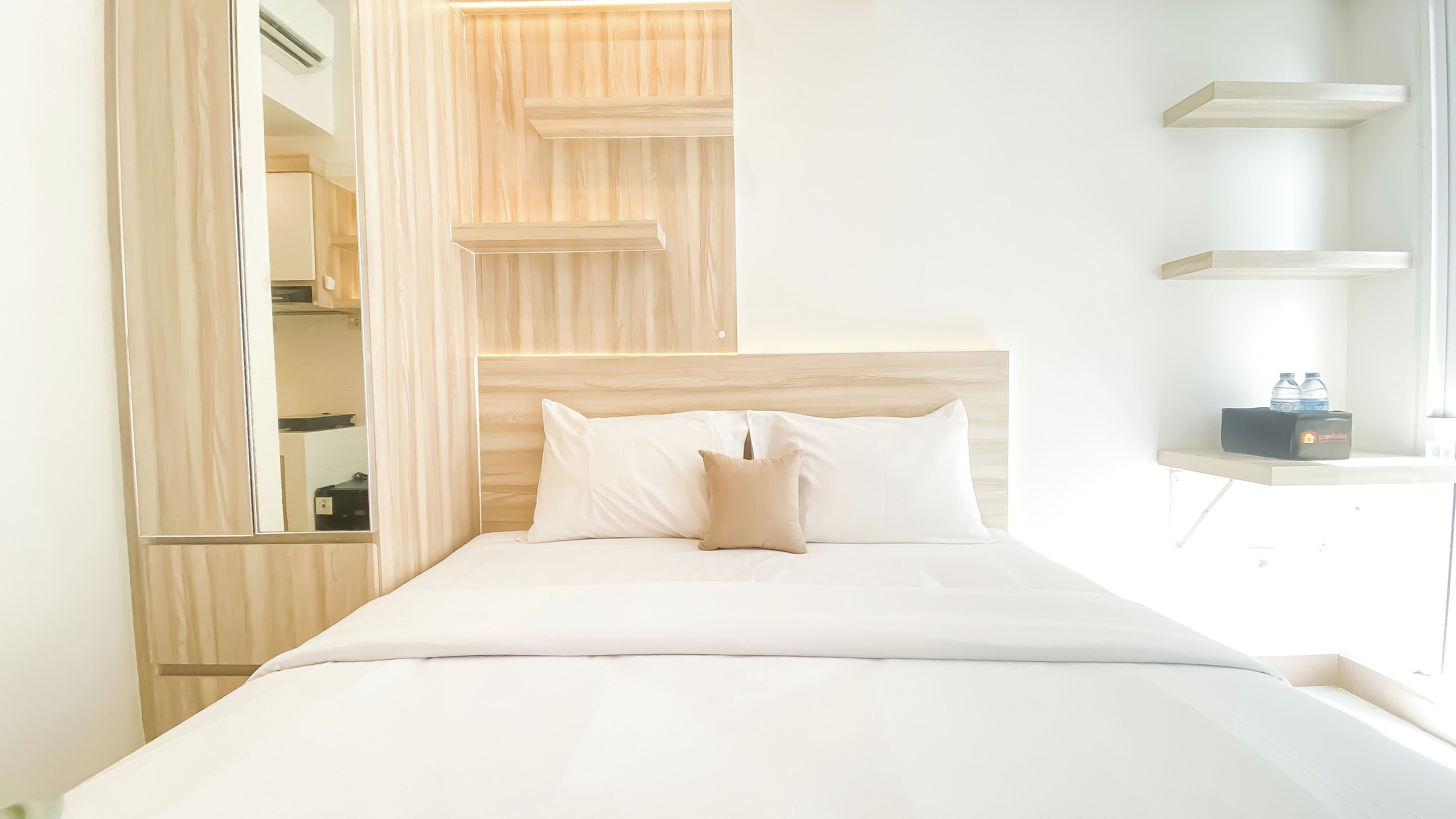 A neatly made bed with white linens and a beige pillow, a wooden headboard, and a small bedside table with a water bottle and a device.