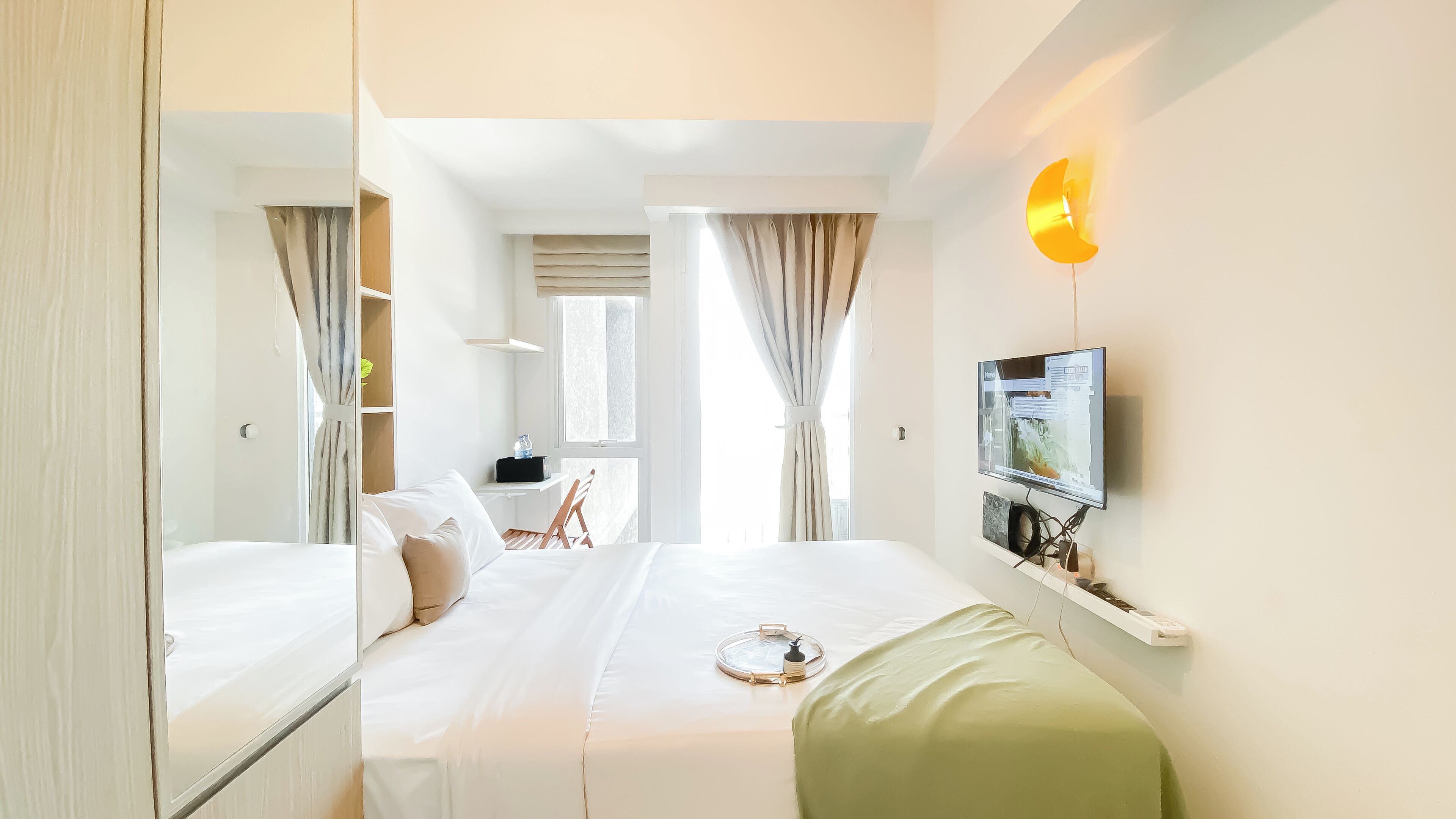 A hotel room with a bed, a desk, a chair, a TV mounted on the wall, and a window with curtains.