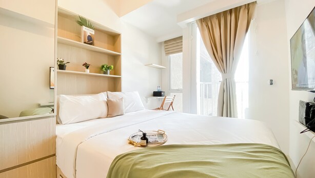 A hotel room with a bed, a bedside table, a shelf with plants, a window with curtains, and a wall-mounted TV. - Cozy Living and Homey Studio at Apartment Tokyo Riverside PIK 2 (Teluknaga)