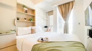 Habitación de hotel con cama, mesita de noche, estante con plantas, ventana con cortinas y televisor montado en la pared.
