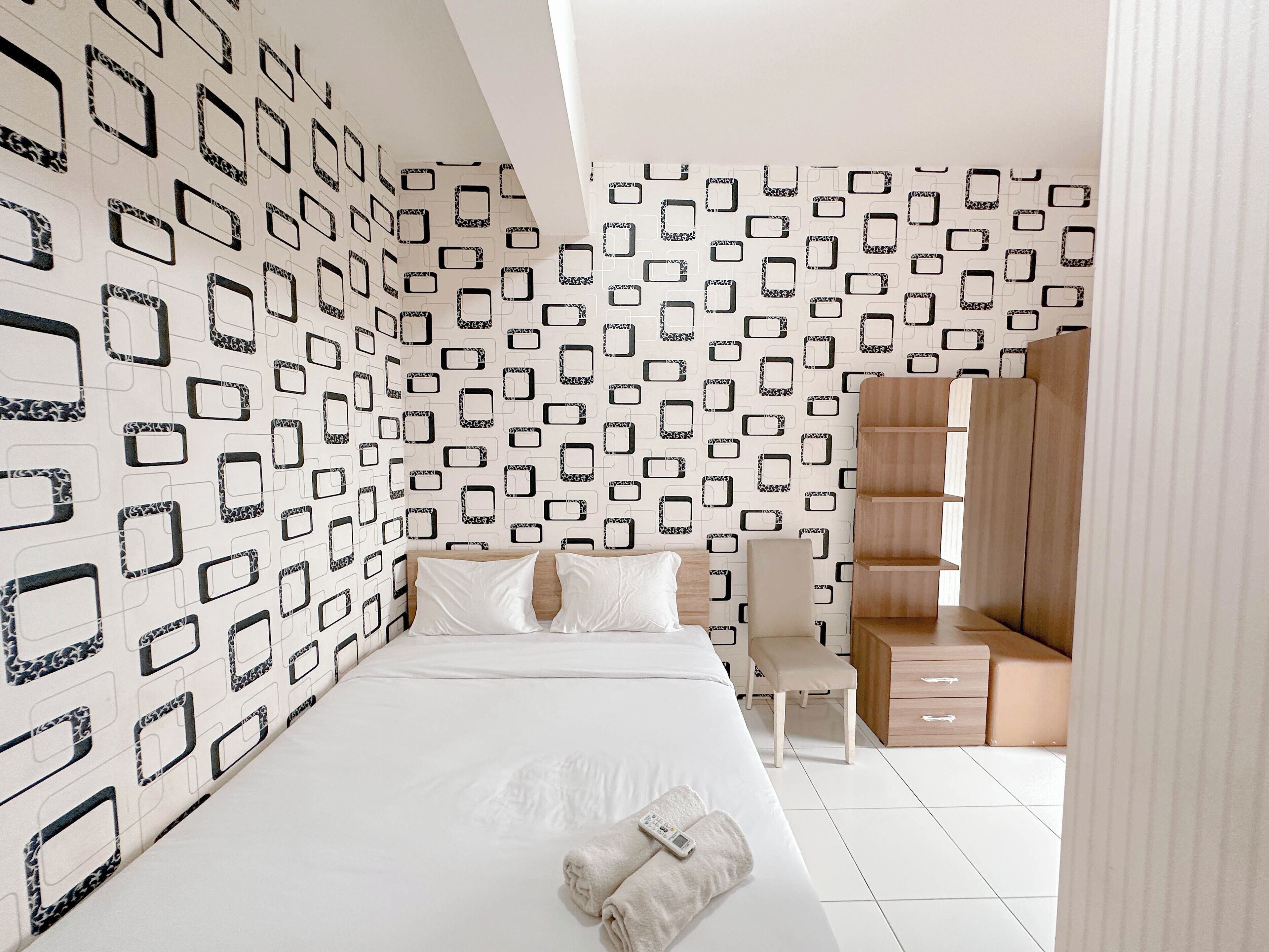 A modern hotel room with a geometric patterned wall, a neatly made bed, a wooden wardrobe, and a small desk.