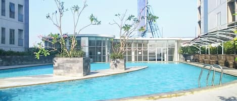 Une piscine dotée d’une enceinte en verre, entourée de plantes en pot et bordée d’un bâtiment moderne sur lequel flotte un drapeau.