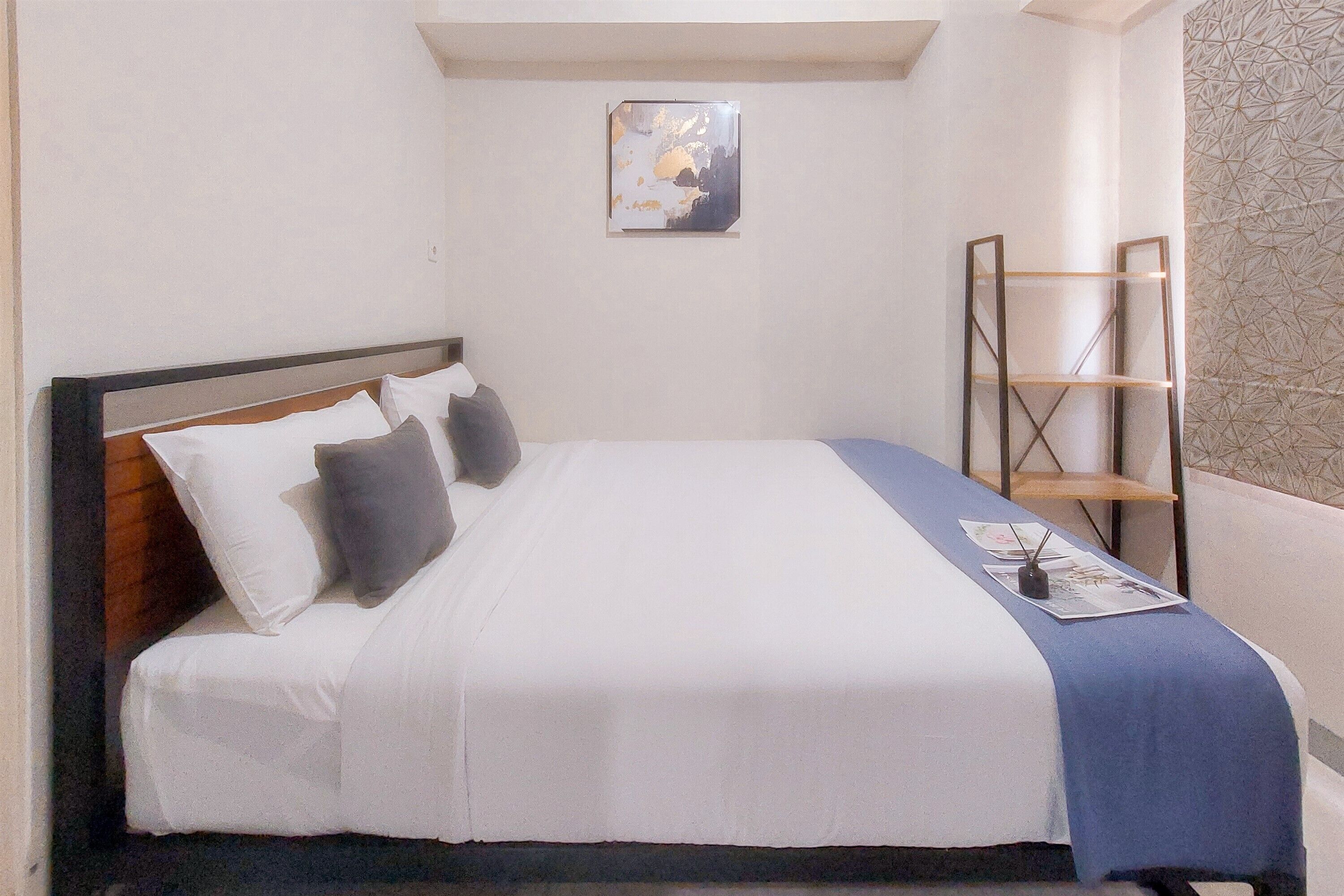 Una cama bien hecha con ropa de cama blanca y gris, una manta azul y una mesita con un libro y un control remoto.