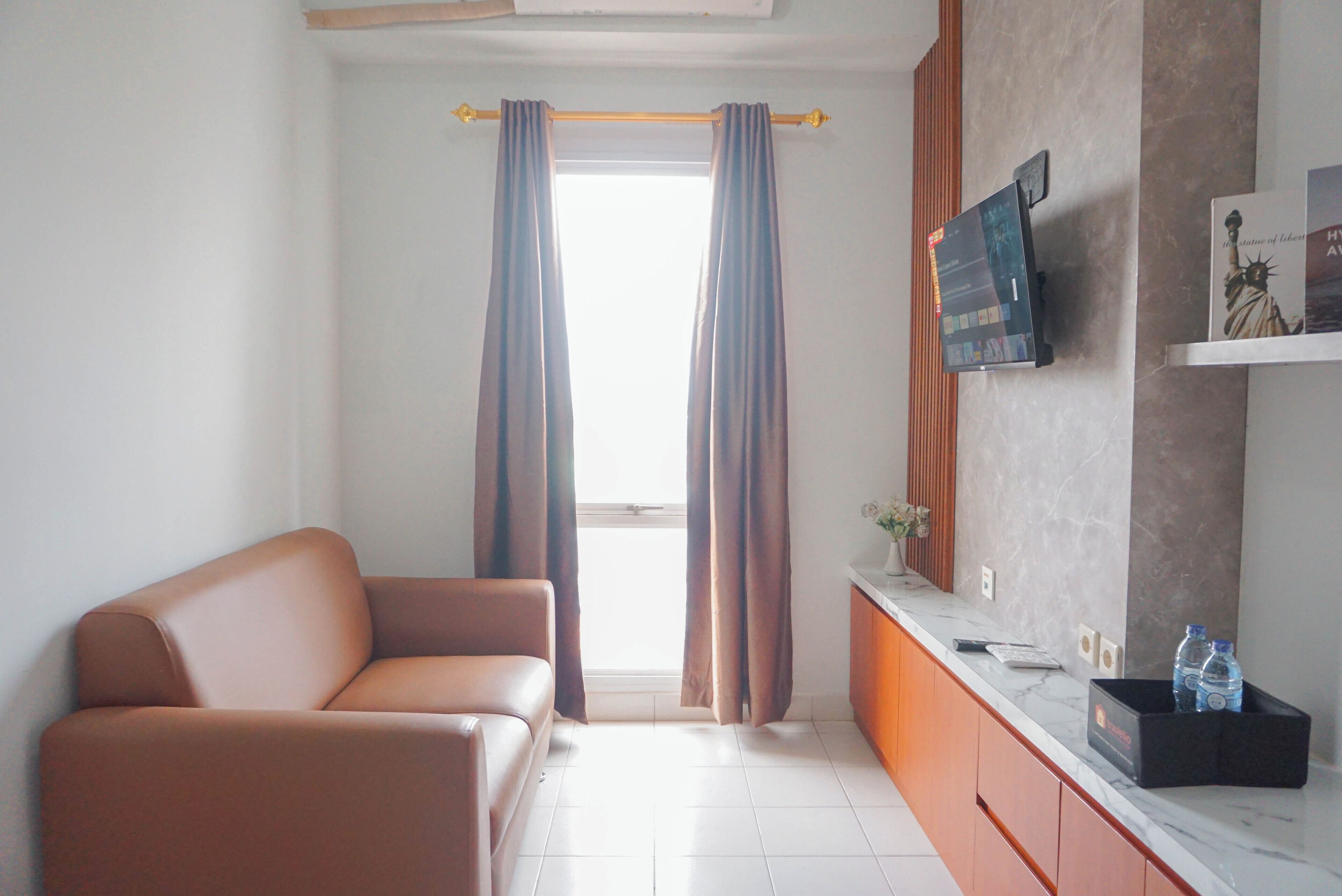 A modern living room with a sofa, a flat-screen TV mounted on the wall, a small table with a vase and a couple of bottles, and a window with curtains.