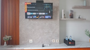 Een hotel receptie met een aan de muur bevestigde televisie waarop een film draait, een wand van houten latten, een marmeren balie, een vaas met bloemen en een plank met boeken en een houder voor waterflessen.
