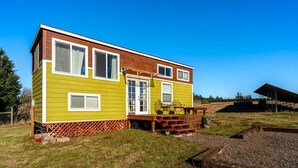 A two-story mobile home with a wooden deck and a small porch, surrounded by a grassy area and a fenced yard.