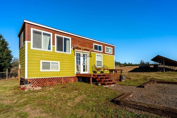 A two-story mobile home with a wooden deck and a small porch, surrounded by a grassy area and a fenced yard.
