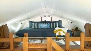 A bedroom with a sloped ceiling, a bed with a blue bedspread, multiple pillows, a wooden headboard, a small bedside table with a lamp, and a window with curtains.