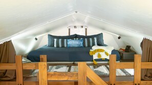 A bedroom with a sloped ceiling, a bed with a blue bedspread, multiple pillows, a wooden headboard, a small bedside table with a lamp, and a window with curtains.