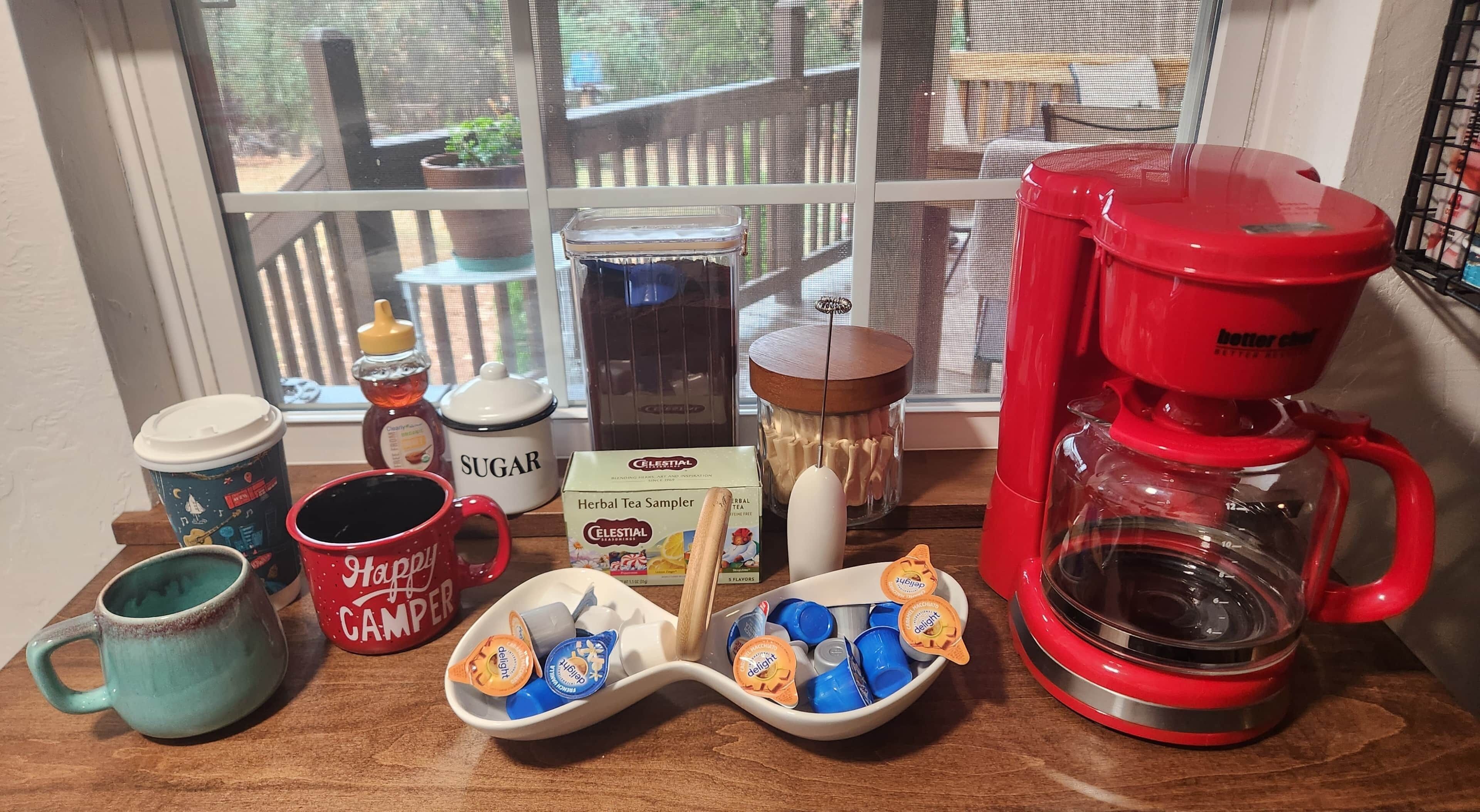 A kitchen counter with a coffee maker, sugar container, tea sampler, and various mugs.