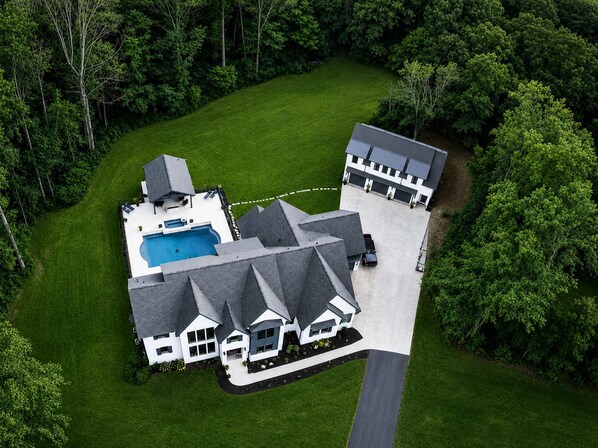 A large house with a swimming pool, surrounded by a well-maintained lawn and dense trees.