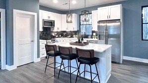 A modern kitchen with white cabinets, a stainless steel refrigerator, a central island with bar stools, and pendant lighting. - Luxury Guest House w/Resort Style Pool & Spa, pets welcome (Huntersville)