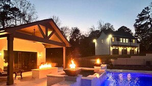 A pool with blue lighting, a covered patio with fire pits, and a house in the background. - Luxury Guest House w/Resort Style Pool & Spa, pets welcome (Huntersville)