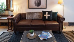 A living room with a brown leather sofa, a chandelier, a framed deer artwork, a floor lamp, and a vintage suitcase with books. - Matilda - Stunning, 3-bed, vintage home in Duluth! (Duluth)