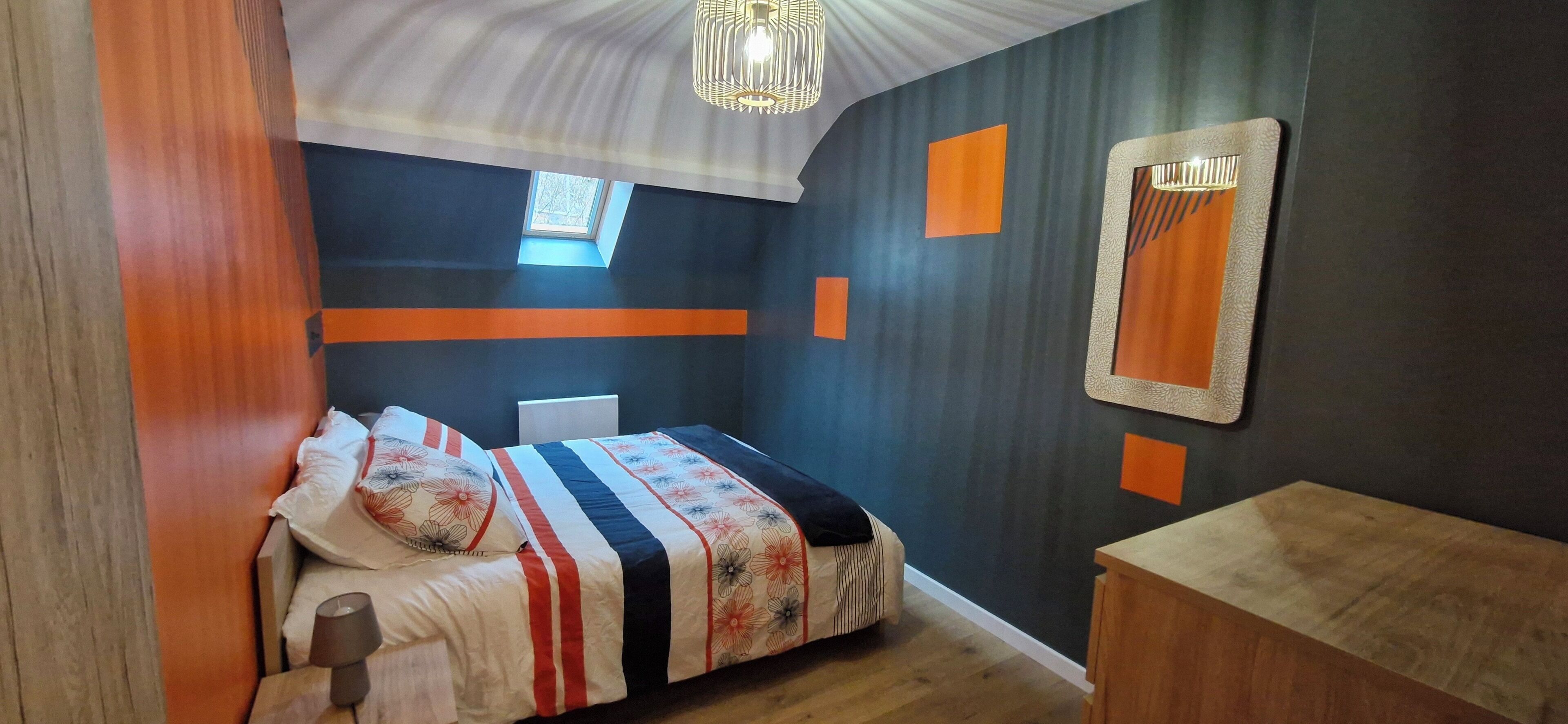 A bedroom with a bed, a nightstand, a wooden dresser, a window, and a mirror on the wall.