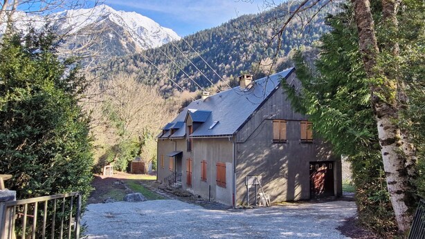 Un bâtiment rustique doté d’un toit en pente, d’une petite remise attenante et d’une allée en gravier menant à une entrée fermée par un portail.