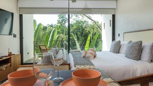 Quarto moderno com uma cama grande, uma mesa de jantar com tigelas e copos de vinho, e uma varanda com vegetação exuberante.