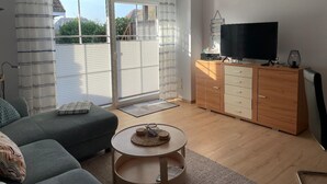 Een woonkamer met een bank, een salontafel, een tv op een houten kast en een glazen schuifdeur naar een balkon.