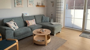 Een moderne woonkamer met een grijze bank, een ronde houten salontafel en uitzicht op een bakstenen gebouw door een groot raam.