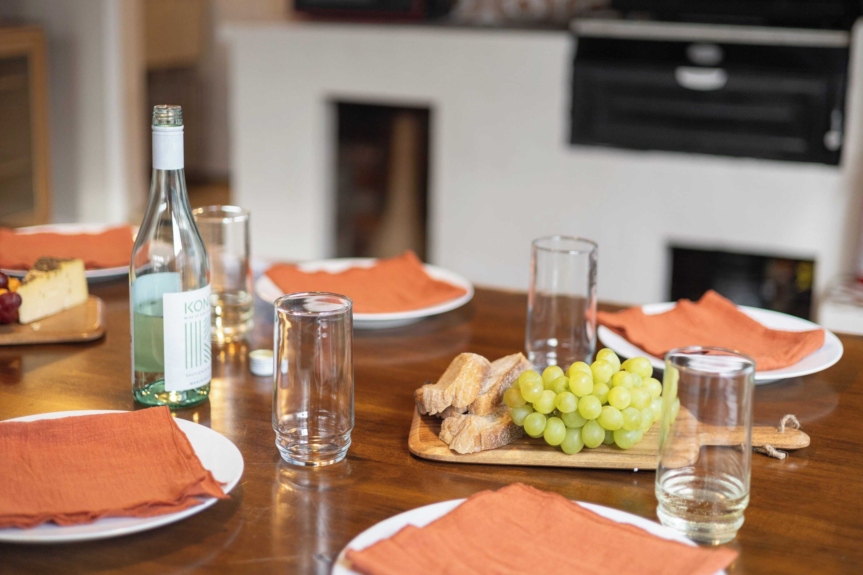 A dining table with plates, a bottle of wine, glasses, and a wooden board with grapes and cheese.
