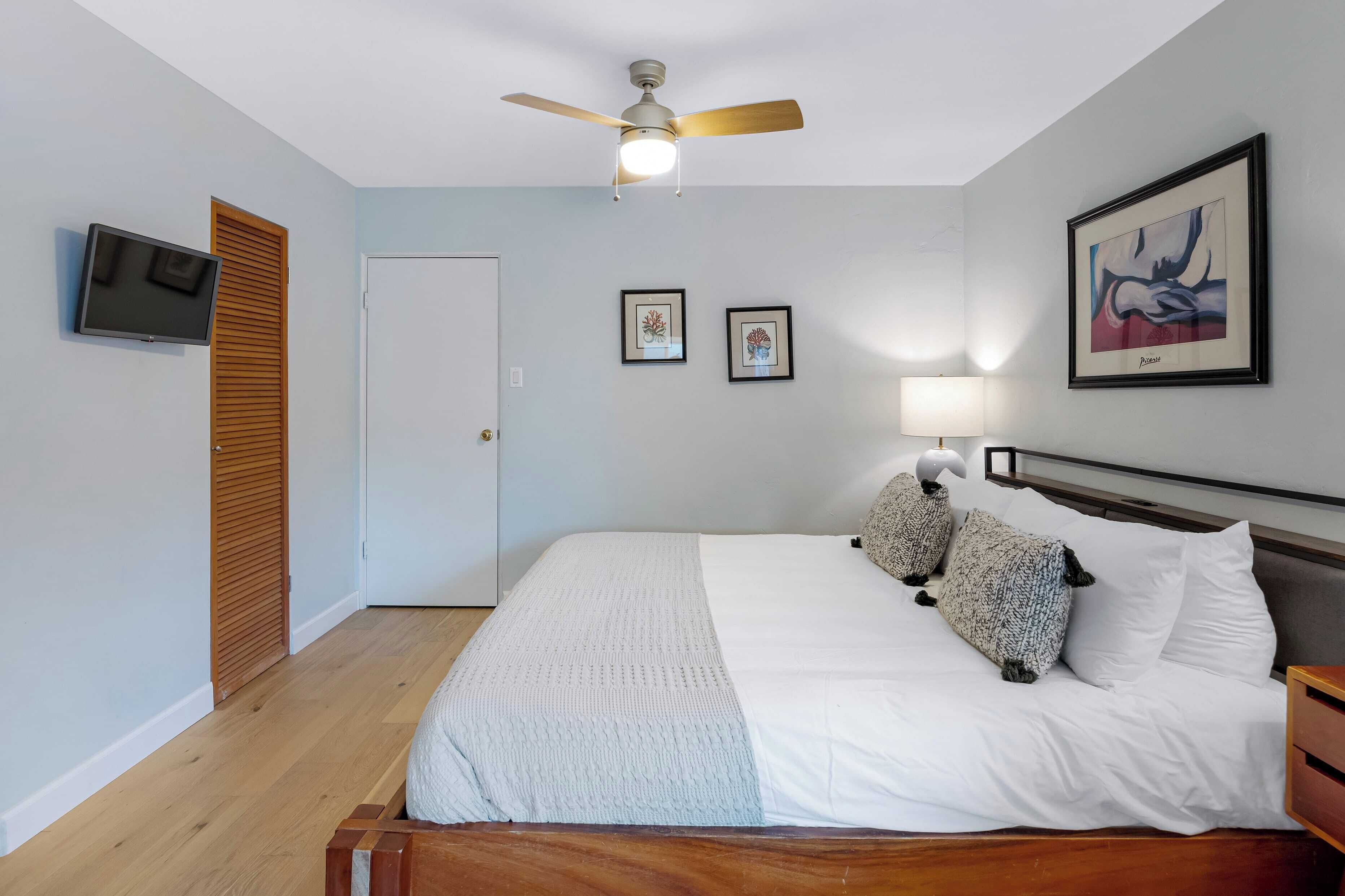 A bedroom with a bed, wooden headboard, ceiling fan, wall-mounted TV, framed artwork, and a door.