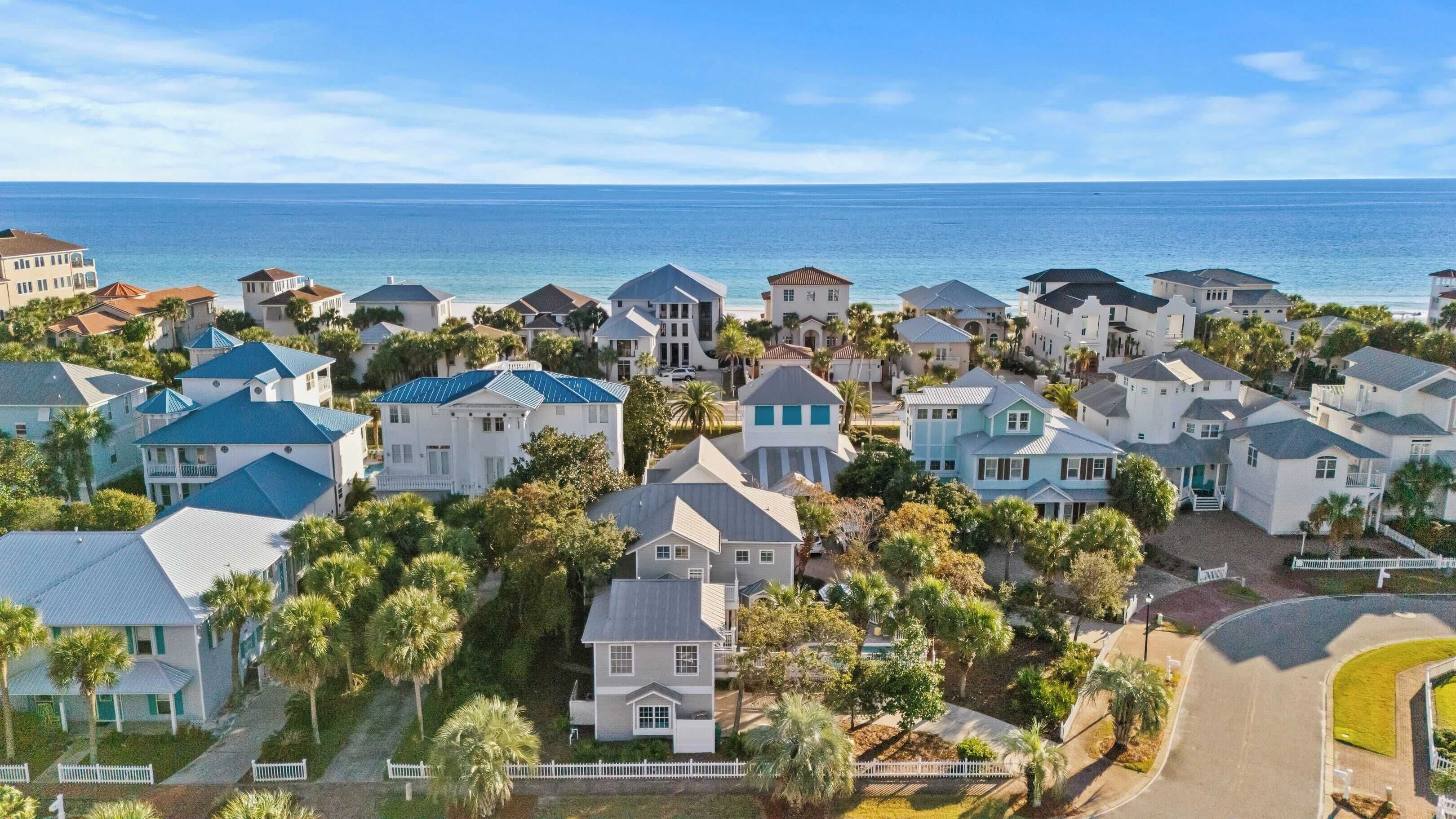 Un quartier résidentiel en bord de mer où les maisons présentent différents styles architecturaux, notamment des toits à pignon et des façades en stuc, et sont entourées de palmiers, offrant une vue sur l’océan.