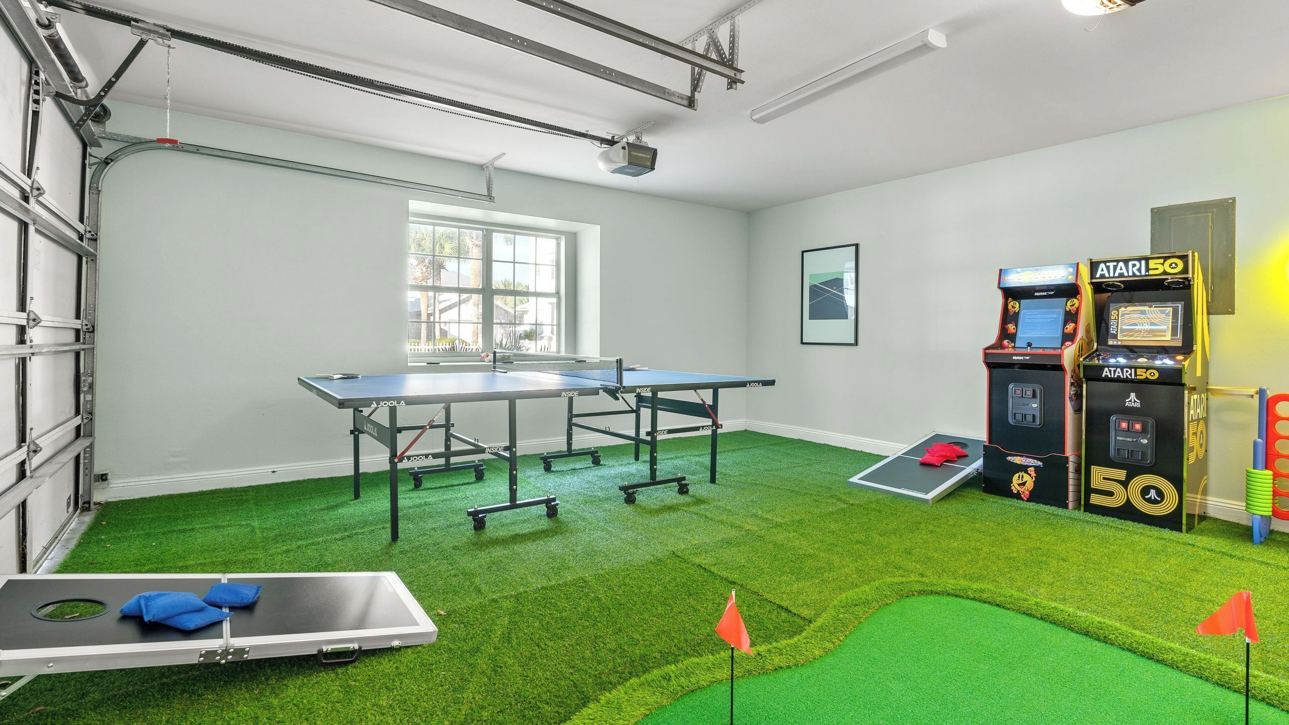A ping pong table, arcade games, and artificial grass in a room with a window and a projector.