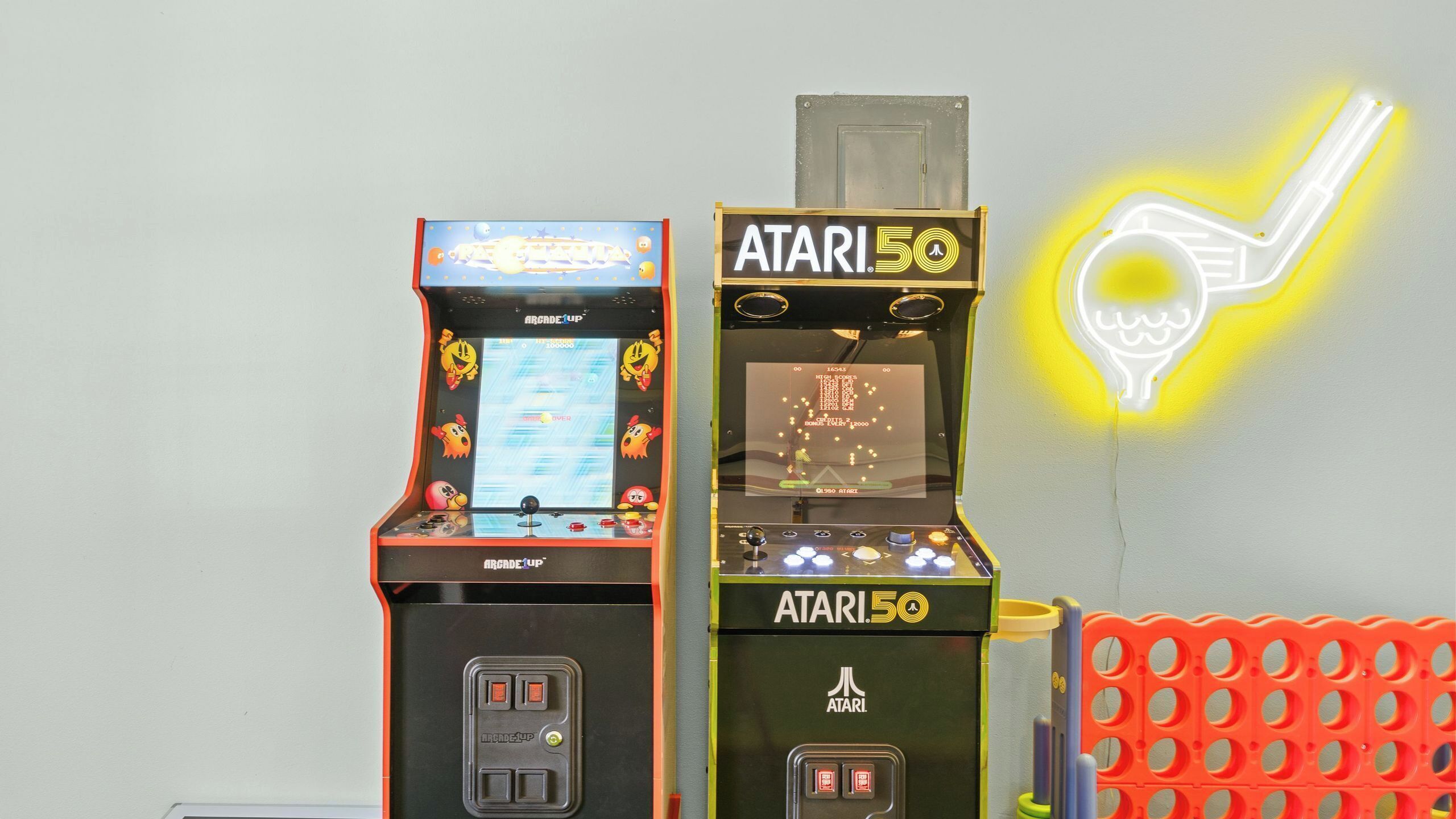 Two arcade machines, one labeled 'Arcade Up' and the other 'Atari 50,' with a neon light decoration in the background.