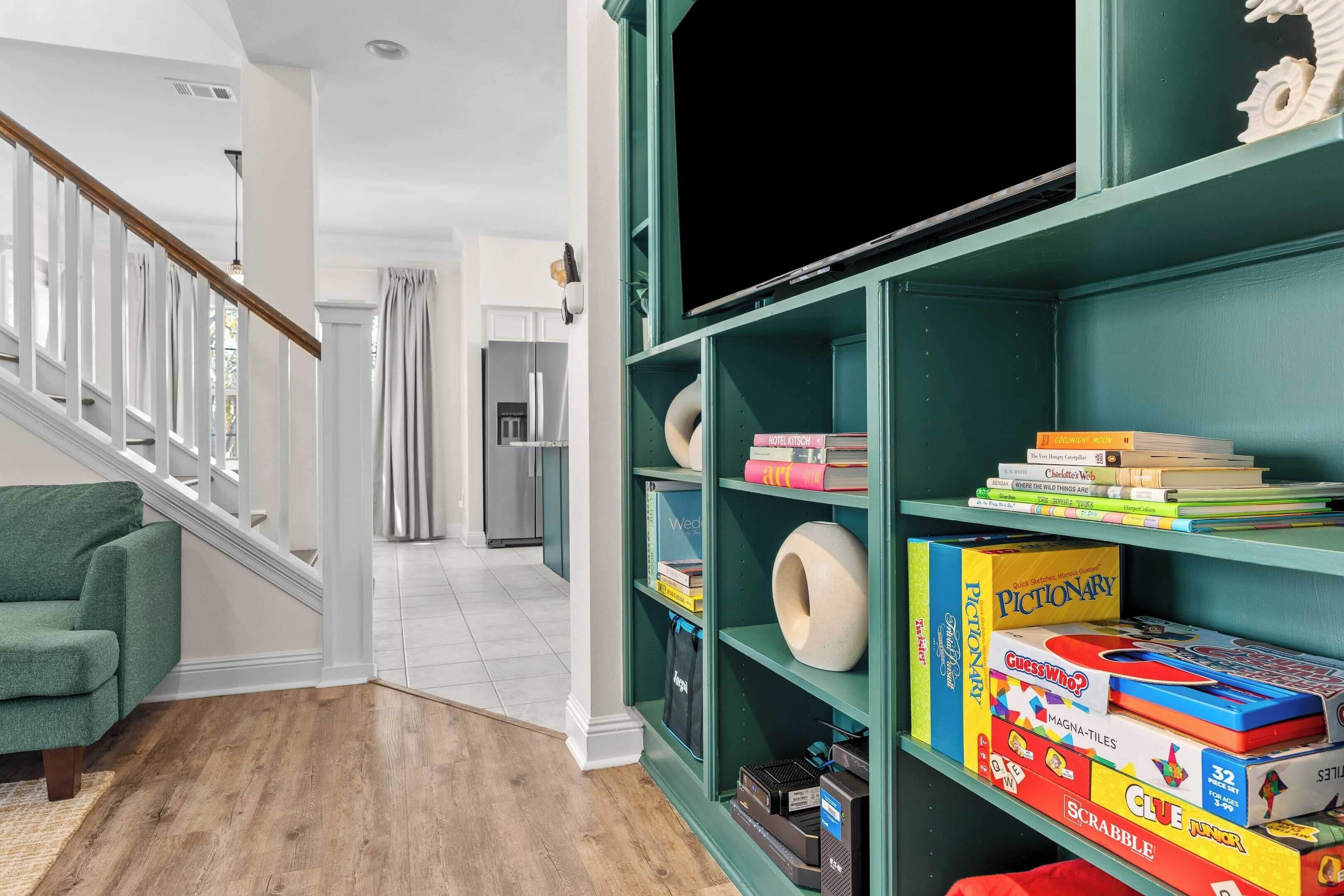Un salon avec un canapé vert, une bibliothèque remplie de jeux de société et un escalier menant à une autre pièce.