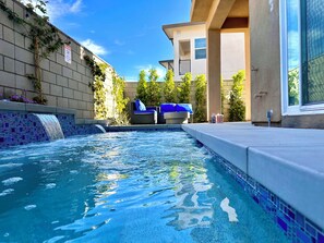 A swimming pool with a blue mosaic tile border, two lounge chairs, and a building with a beige facade in the background.