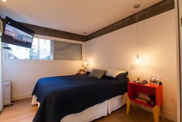 A bedroom with a bed, a nightstand, a red side table, a window with blinds, and a TV mounted on the wall. - Spacious, modern, and cozy apartment in a great location (Itaim Bibi São Paulo)