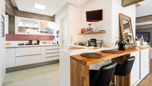 A modern kitchen with white cabinets, a wooden countertop, and a built-in stove. There is a dining area with a wooden table and black chairs. A flat-screen TV is mounted on the wall above the counter. - Spacious, modern and cozy apartment very well located (Itaim Bibi São Paulo)