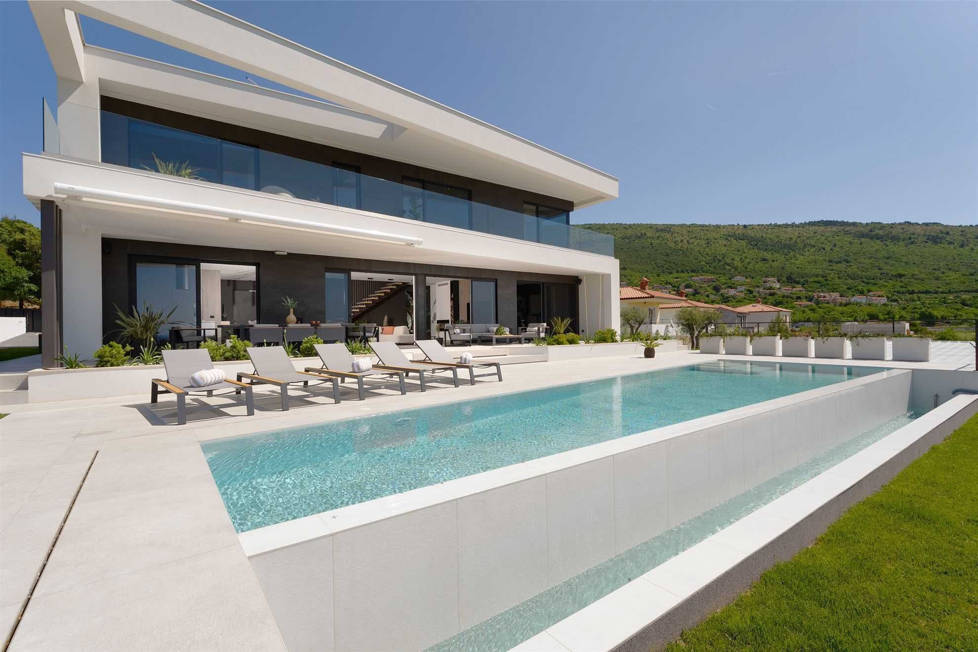 A modern two-story house with a large swimming pool, outdoor seating, and a view of a green hillside.