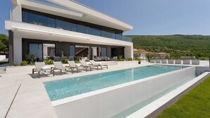 A modern two-story house with a large swimming pool, outdoor seating, and a view of a green hillside.