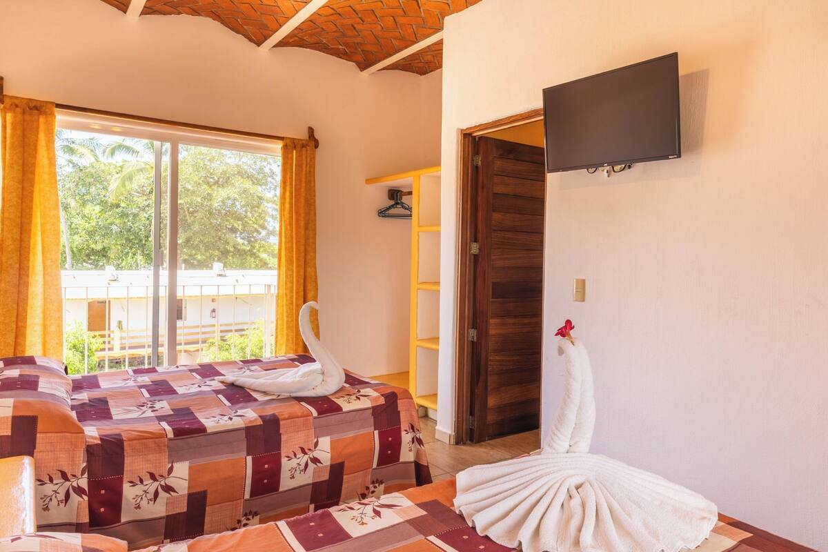 Una habitación con una cama, un televisor fijado a la pared y un ventanal con cortinas.