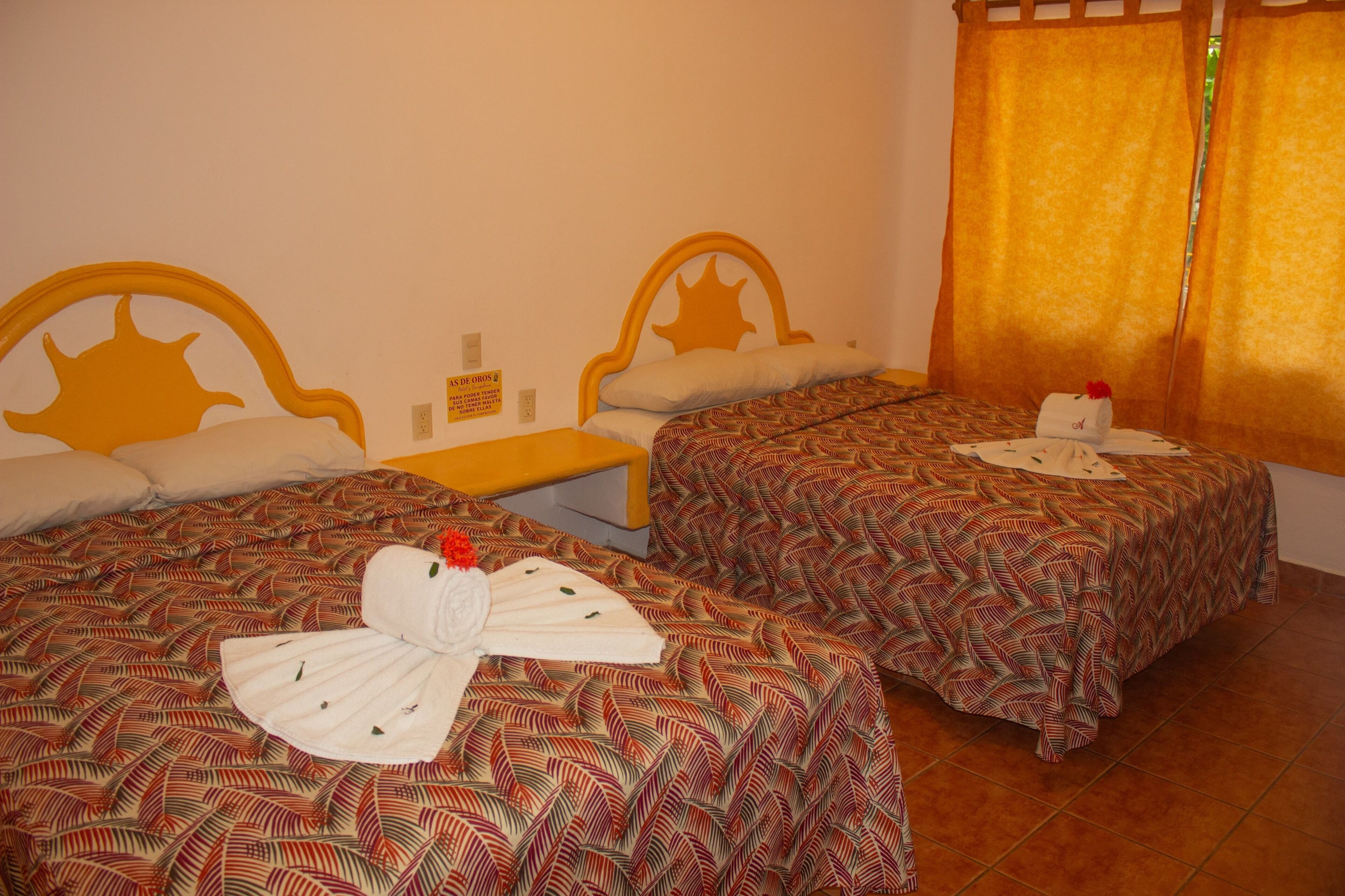 Habitación de hotel con dos camas, cada una con una colcha estampada y cabeceros con un diseño que recuerda al sol. Hay una mesita entre las camas y un cartel en la pared.