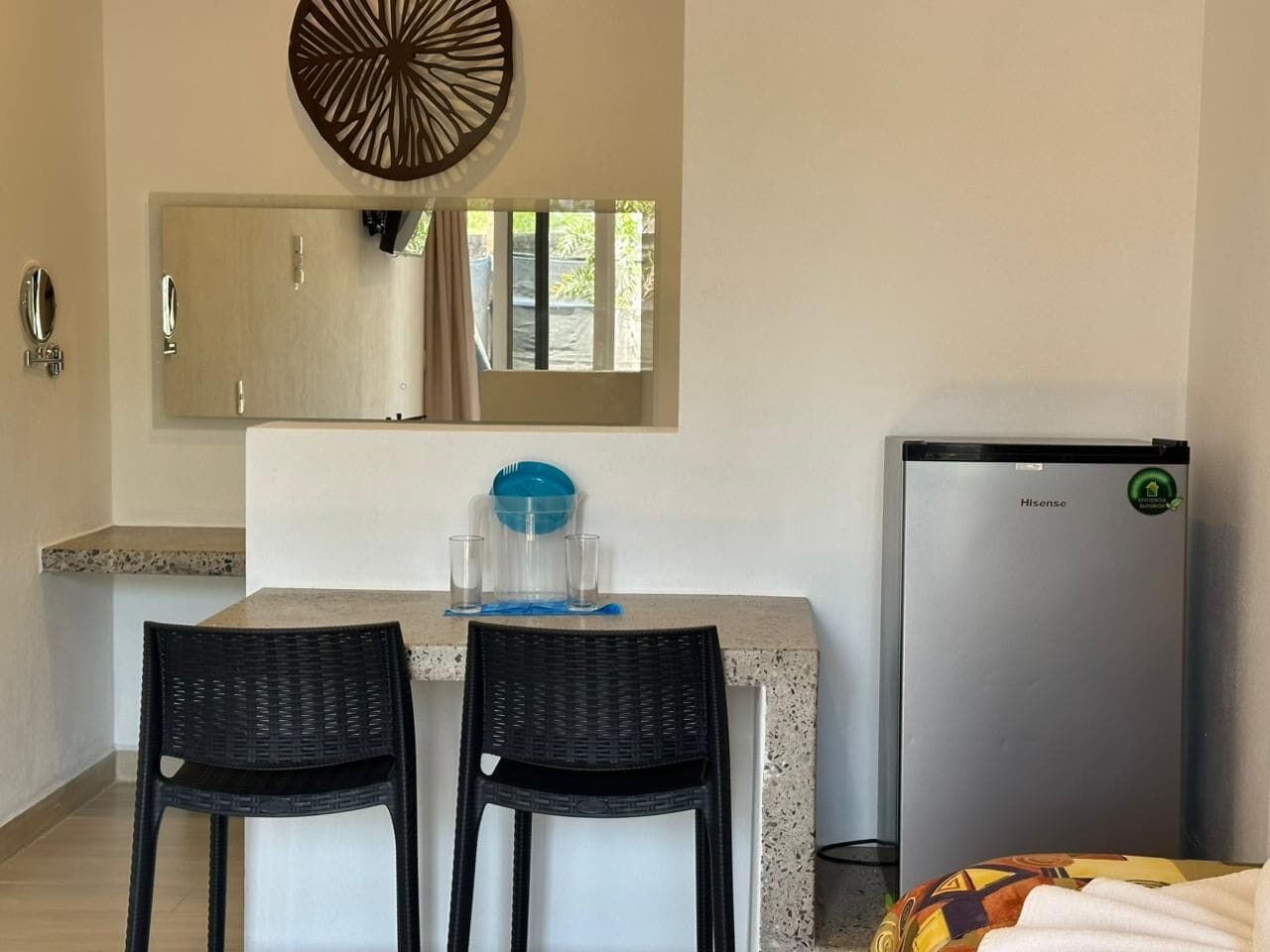 A compact kitchen with a refrigerator, two chairs, a dining table, and a mirror above the counter.