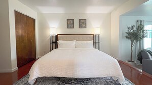 Une chambre avec un grand lit, une porte en bois, deux tableaux accrochés au mur et une plante en pot.