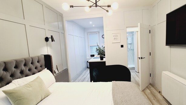 Un dormitorio con cabecera capitonada, cama con ropa de cama blanca y beige, escritorio con silla y televisor montado en la pared.