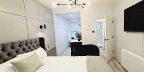 Ein Schlafzimmer mit einer gesteppten Kopfteil, einem Bett mit weißer und beiger Bettwäsche, einem Schreibtisch mit Stuhl sowie einem an der Wand befestigten Fernseher.