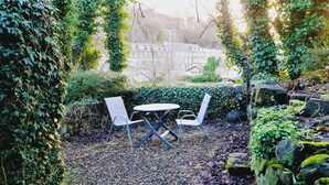 Un área de estar al aire libre con dos sillas y una mesa, rodeada de abundante follaje y rocas.