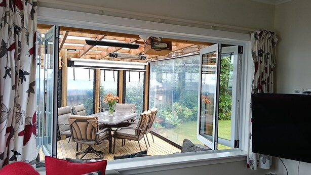 Een kamer met een grote schuifglazen deur die uitkomt op een terras buiten. Binnenin staat een bank met rode en blauwe kussens, een televisie en een houten kruk.