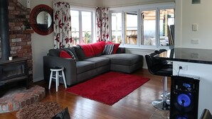 Een woonkamer met een open haard, een grijze bank met rode en zwarte kussens, een rode vloerkleed en een computerbureau met een monitor en toetsenbord.