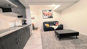 A modern kitchen with dark cabinets and a countertop, a black sofa with colorful cushions, and a small coffee table. - Luxury and Modern 2 bedroom suite (Balzac)