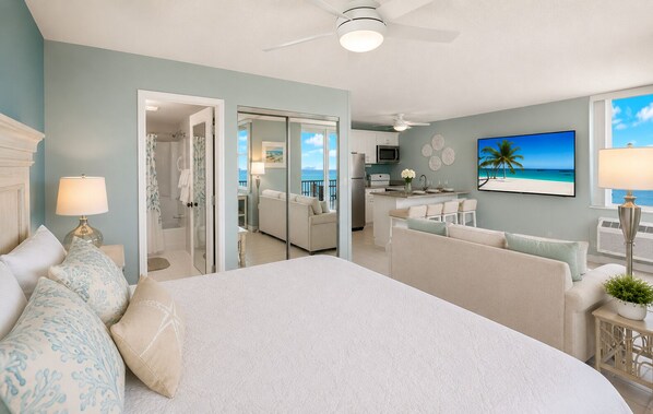 A bedroom with a large bed, a sitting area with a sofa, a dining area with a table and chairs, and a view of the ocean through a glass door.
