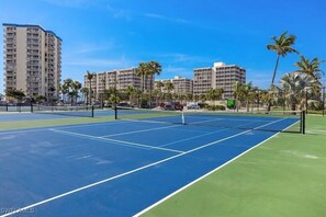 A tennis court with a net, surrounded by palm trees and buildings.