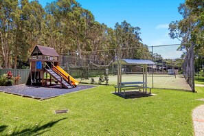 Un parque infantil con un tobogán y una casita, rodeado de una cerca y árboles.