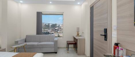 Une chambre d’hôtel moderne avec un lit, un canapé gris, une petite table, un bureau et une vue sur la ville par la fenêtre.