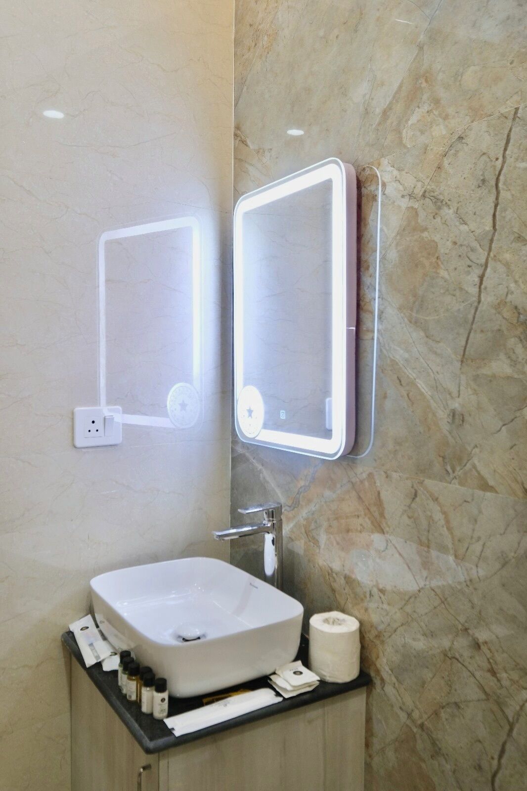 A bathroom with a marble wall, a white sink, and two illuminated mirrors.